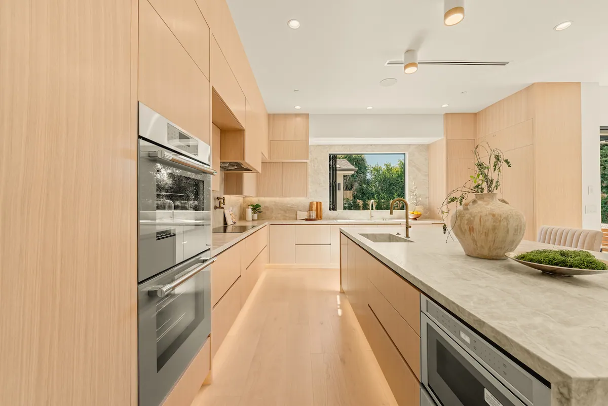 Remodeled galley kitchen with white oak cabinetry, stacked wall ovens and a stone-topped island